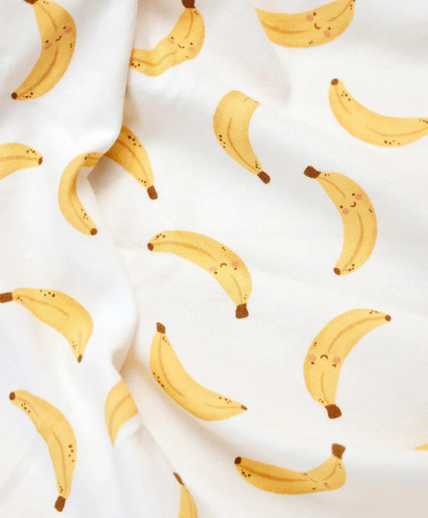 Organic cotton baby swaddle wrap featuring a playful banana print in warm yellow tones on a white fabric