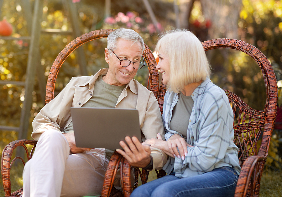 Happy elderly couple relaxing in wicker chairs in open air and using laptop computer,